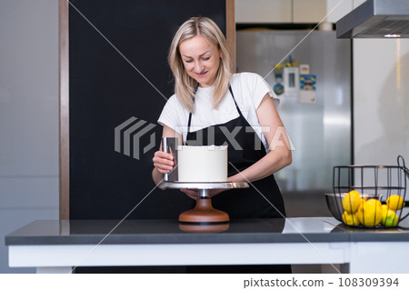 Happy blonde woman finishing processing white mastic on sugar free cake with pastry scraper self-made confectioner enjoying preparing custom confectionery for anniversary Happy blonde woman finishing processing white mastic on sugar free cake with pastry scraper self-made confectioner enjoying preparing custom confectionery for anniversary 108309394