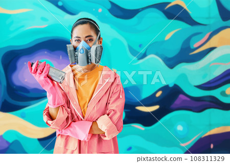 Graffiti female street artist in respirator mask standing near the wall with her paintings looking to camera Graffiti female street artist in respirator mask standing near the wall with her paintings looking to camera 108311329