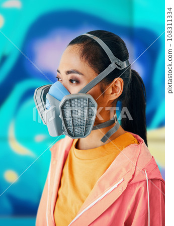 Close up portrait of graffiti street artist in respirator mask standing near the wall with her colorful paintings 108311334