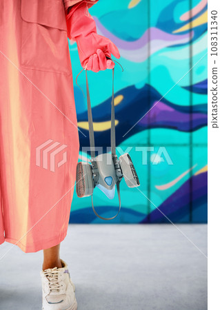 Cropped image of street artist with respirator mask in hand standing near the wall with her paintings 108311340