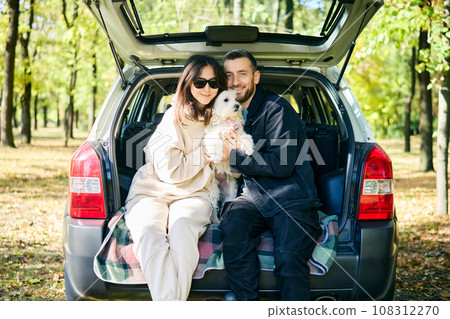 Loving couple hugging their fluffy white dog while relaxing in car trunk in forest Loving couple hugging their fluffy white dog while relaxing in car trunk in forest 108312270