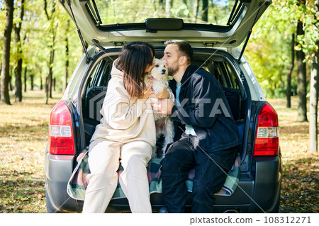 Couple affectionately kissing their lovely dog sitting in car trunk relaxing in the forest Couple affectionately kissing their lovely dog sitting in car trunk relaxing in the forest 108312271