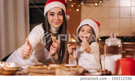 Adorable little girl and her mom in santa hat eating homemade gingerbread and laughing. Happy little kid preparing for Christmas. Background of fairy lights bokeh 108312586