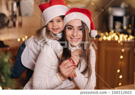 Portrait of adorable little daughter with her mom at kitchen during Christmas holidays. Girls in santa hat. Decorative fit tree at the table. Girls make gingerbread Portrait of adorable little daughter with her mom at kitchen during Christmas holidays. Girls in santa hat. Decorative fit tree at the table. Girls make gingerbread 108312603