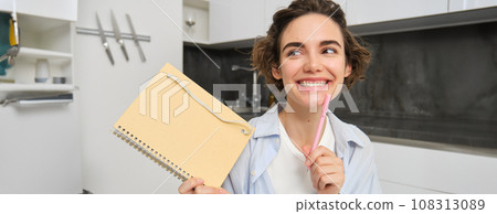 Portrait of young creative woman, holding her notebook and smiling, makes notes in her diary, writing in her planner, sitting at home in kitchen and smiling 108313089