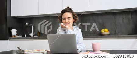 Young female entrepreneur works from homes. Girl with laptop sits in her kitchen, does homework, works on remote, connects to online meeting, taking notes Young female entrepreneur works from homes. Girl with laptop sits in her kitchen, does homework, works on remote, connects to online meeting, taking notes 108313090