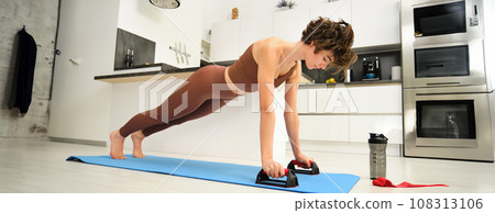 Young fitness woman, stands on push up bars stand, doing plunk exercise at home, does chest press workout training in her kitchen, exercising on yoga rubber mat 108313106