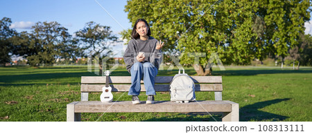 Girl with sad face, sitting on bench with smartphone and ukulele, looking upset and disappointed, being alone in park 108313111