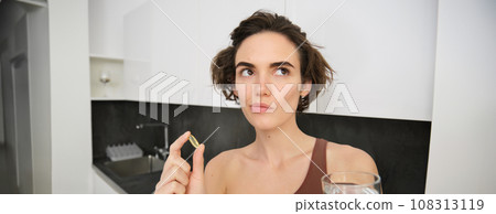 Close up portrait of sportswoman in activewear, holding omega-3, vitamin c, d and b in her hand, taking dietary supplement with drinking glass of water. Healthy lifestyle and sport concept Close up portrait of sportswoman in activewear, holding omega-3, vitamin c, d and b in her hand, taking dietary supplement with drinking glass of water. Healthy lifestyle and sport concept 108313119
