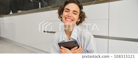 Portrait of young adult woman works on her tablet, sits with notebook on kitchen floor at home, studies in comfort Portrait of young adult woman works on her tablet, sits with notebook on kitchen floor at home, studies in comfort 108313121
