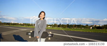Vertical shot of skater girl posing with longboard, cruising on empty road in suburbs. Smiling asian woman skating on skateboard, holding cruiser in hands 108313130