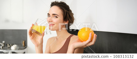 Image of sportswoman, fitness girl holding glass of juice and an orange, smiling, drinking vitamin beverage after workout, standing in her kitchen at home. Healthy lifestyle and sport concept Image of sportswoman, fitness girl holding glass of juice and an orange, smiling, drinking vitamin beverage after workout, standing in her kitchen at home. Healthy lifestyle and sport concept 108313132