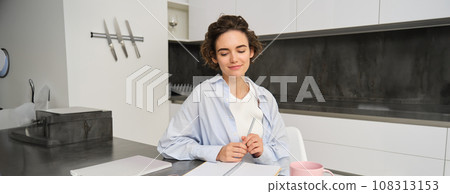 Portrait of young woman working from home, writing down information in notebook, taking notes, sitting in kitchen and studying, student doing homework Portrait of young woman working from home, writing down information in notebook, taking notes, sitting in kitchen and studying, student doing homework 108313153