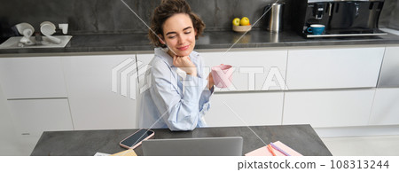 Beautiful woman managing her work from home, drinking coffee and looking at laptop, sitting in kitchen with paperwork, doing her taxes, studying indoors on remote Beautiful woman managing her work from home, drinking coffee and looking at laptop, sitting in kitchen with paperwork, doing her taxes, studying indoors on remote 108313244