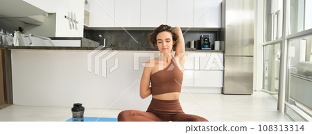 Portrait of brunette girl doing sports at home, sitting on yoga mat in bright room at home, stretching hands behind her back, workout indoors 108313314