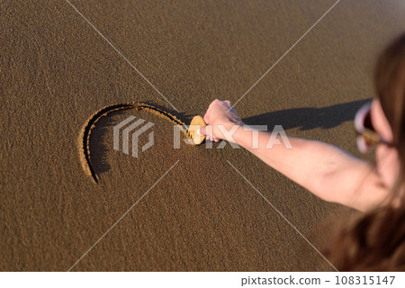 Young woman on the beach drawing a heart shape on the sand at sunset. Young woman on the beach drawing a heart shape on the sand at sunset. 108315147