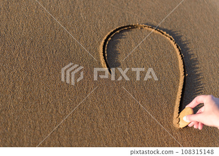 Young woman on the beach drawing a heart shape on the sand at sunset. Young woman on the beach drawing a heart shape on the sand at sunset. 108315148