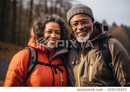 Portrait of senior African American couple in touristic outerwear with backpacks. Elderly active man and woman hiking and walking along autumn forest. Healthy lifestyle for retired people. Portrait of senior African American couple in touristic outerwear with backpacks. Elderly active man and woman hiking and walking along autumn forest. Healthy lifestyle for retired people. 108316835