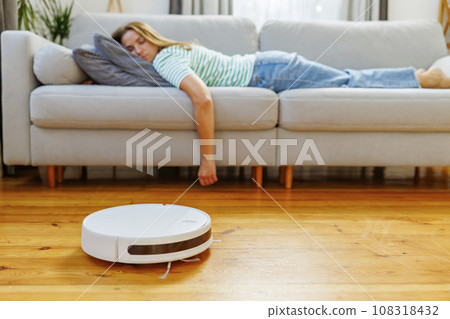 A woman sleeping on sofa while robot vacuum quietly takes care of the cleaning 108318432