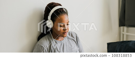 Banner smiling young african woman listening to music with headphones while relaxing on bed at home, meditating. Copy space and empty space for text 108318853