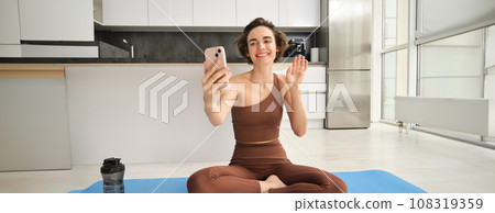 Smiling fitness girl, young athlete sits at home on yoga mat, waves hand at smartphone, joing online workout training session from home, says hello Smiling fitness girl, young athlete sits at home on yoga mat, waves hand at smartphone, joing online workout training session from home, says hello 108319359