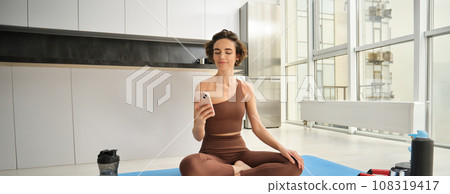 Sport app and wellbeing. Young woman, athlete sitting on floor at home with smartphone, doing yoga, using workout app on mobile phone, doing exercises at home 108319417