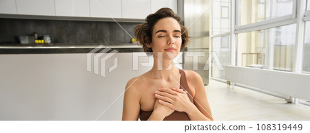 Portrait of young mindful woman, opens her chakra, doing yoga on rubber mat at home, closes eyes and smiles, feels peace inside her chest. Portrait of young mindful woman, opens her chakra, doing yoga on rubber mat at home, closes eyes and smiles, feels peace inside her chest. 108319449