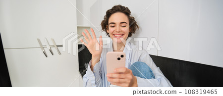 Happy smiling girl, saying hi to a friend on video chat, waves at smartphone app, connects to online conversation on her phone 108319465