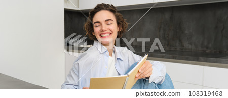 Portrait of happy smiling woman, holding notebook, studying in her kitchen, reading journal 108319468