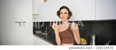 Portrait of sportswoman drinking water, taking vitamins, dietary supplements for healthy skin, having omega-3 pill, standing in her kitchen in workout clothing Portrait of sportswoman drinking water, taking vitamins, dietary supplements for healthy skin, having omega-3 pill, standing in her kitchen in workout clothing 108319470