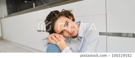 Close up portrait of beautiful, tender young woman smiling, looking at camera, sitting in cozy pose on kitchen floor, daydreaming 108319582