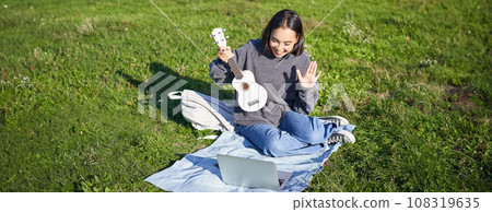 Cheerful asian girl, musician waves at her laptop, say hi to video chat, plays ukulele and records it on laptop, sits on grass in park 108319635
