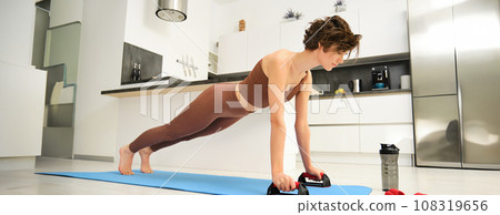 Portrait of young woman in sportswear, workout on yoga rubber mat, stands on push up bars stand for plunk exercises, fitness training session at home Portrait of young woman in sportswear, workout on yoga rubber mat, stands on push up bars stand for plunk exercises, fitness training session at home 108319656