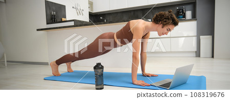 Portrait of young sportswoman in activewear, standing in plunk, following online video gym class on laptop, drinking water during her workout training session at home, exercising in kitchen Portrait of young sportswoman in activewear, standing in plunk, following online video gym class on laptop, drinking water during her workout training session at home, exercising in kitchen 108319676