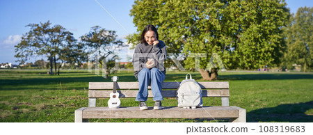 Excited girl looks at smartphone and celebrates, wins on mobile phone, sits with ukulele and backpack in park on bench on sunny day 108319683