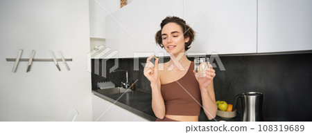 Dietary supplements and healthy lifestyle. Young woman taking vitamin C, D omega-3 with glass of water, standing in activewear, drinking after workout training in her kitchen Dietary supplements and healthy lifestyle. Young woman taking vitamin C, D omega-3 with glass of water, standing in activewear, drinking after workout training in her kitchen 108319689
