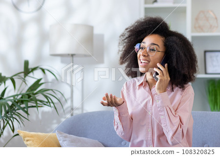 Happy and successful satisfied joyful woman sitting at home on sofa in living room of house talking cheerfully with friends using phone. 108320825