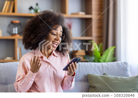 Happy and successful woman winner got good achievement result, african american woman sitting on sofa in living room at home, celebrating success and victory, reading result on smartphone. 108320846