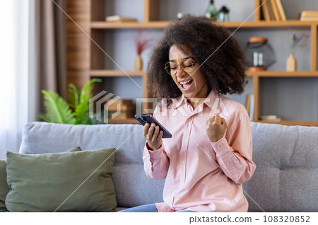 Happy and successful woman winner got good achievement result, african american woman sitting on sofa in living room at home, celebrating success and victory, reading result on smartphone. 108320852