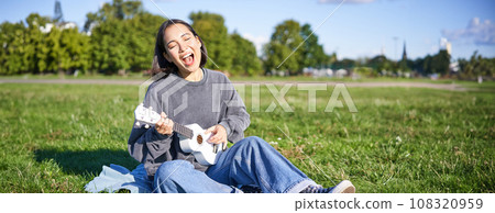 Portrait of beautiful asian woman singing, playing ukulele guitar in park, sitting alone on grass on sunny day Portrait of beautiful asian woman singing, playing ukulele guitar in park, sitting alone on grass on sunny day 108320959