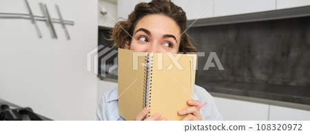 Portrait of cute brunette girl, looks away, hides her face behind planner, notebook, gets creative and writes in her journal 108320972