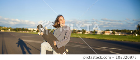 Funny asian girl enjoying skating, holding skateboard like guitar and shadow playing, having fun outdoors Funny asian girl enjoying skating, holding skateboard like guitar and shadow playing, having fun outdoors 108320975
