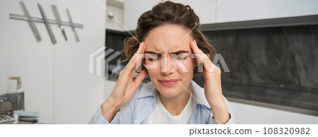Portrait of woman with mgiraine. Girl grimaces from painful headache, touches her head, frowns from discomfort, sits at home 108320982