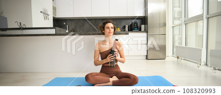 Young strong sporty athletic fitness trainer, instructor woman wearing brown tracksuit sitting on yoga mat drink water training do exercises at home gym indoor. Workout sport motivation concept Young strong sporty athletic fitness trainer, instructor woman wearing brown tracksuit sitting on yoga mat drink water training do exercises at home gym indoor. Workout sport motivation concept 108320993