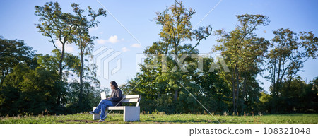 Side view of young woman sitting alone in park on bench, using her laptop to study or work remotely from outdoors Side view of young woman sitting alone in park on bench, using her laptop to study or work remotely from outdoors 108321048