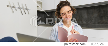 Young woman checking her schedule while working on remote from home, using laptop, looking at her daily planner, making notes, writing down information in notebook 108321269