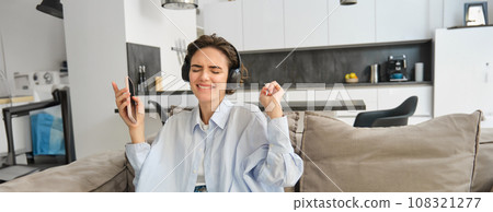 Portrait of beautiful, happy young woman, listening music in headphones, holding smartphone and dancing on sofa at home, enjoys spending time indoors alone Portrait of beautiful, happy young woman, listening music in headphones, holding smartphone and dancing on sofa at home, enjoys spending time indoors alone 108321277