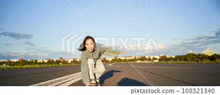 Skater girl riding on skateboard, standing on her longboard and laughing, riding cruiser on an empty street towards the sun Skater girl riding on skateboard, standing on her longboard and laughing, riding cruiser on an empty street towards the sun 108321340