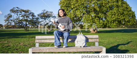 Portrait of young hipster girl sits on bench and plays ukulele, sings along, relaxing in park with her musical instrument Portrait of young hipster girl sits on bench and plays ukulele, sings along, relaxing in park with her musical instrument 108321398