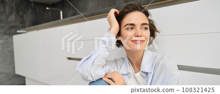 Portrait of young woman sits on floor, smiles and looks outside window, enjoys leisure at home, relaxes, has dreamy face expression Portrait of young woman sits on floor, smiles and looks outside window, enjoys leisure at home, relaxes, has dreamy face expression 108321425